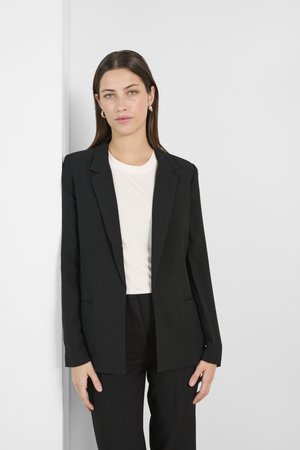 Černý blazer s úzkým střihem, jedno tlačítko na zapínání a dvě přední kapsy. Nesený přes světle zbarvený top. Hladká textilie.