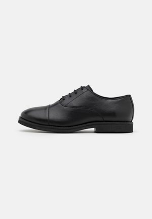 Zapato Oxford de cuero negro para hombre, con cordones cerrados, puntera con costura y tacón bajo apilado, sobre un fondo blanco liso.