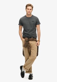 Grå kortærmet t-shirt med en subtil tekstur, khaki bukser og en ternet skjorte bundet om taljen; sorte loafers og ankelsokker.