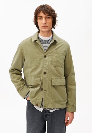 Giovane uomo che indossa una giacca verde oliva con bottoni sopra un maglione lavorato a maglia bianco e nero e jeans scuri, in piedi contro uno sfondo bianco semplice.