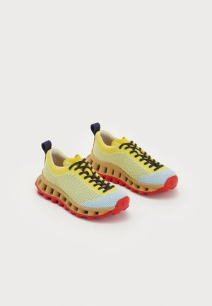 Paar gele en lichtblauwe gebreide sneakers met zwarte veters, beige geventileerde tussenzolen en rode buitenzolen op een witte achtergrond.