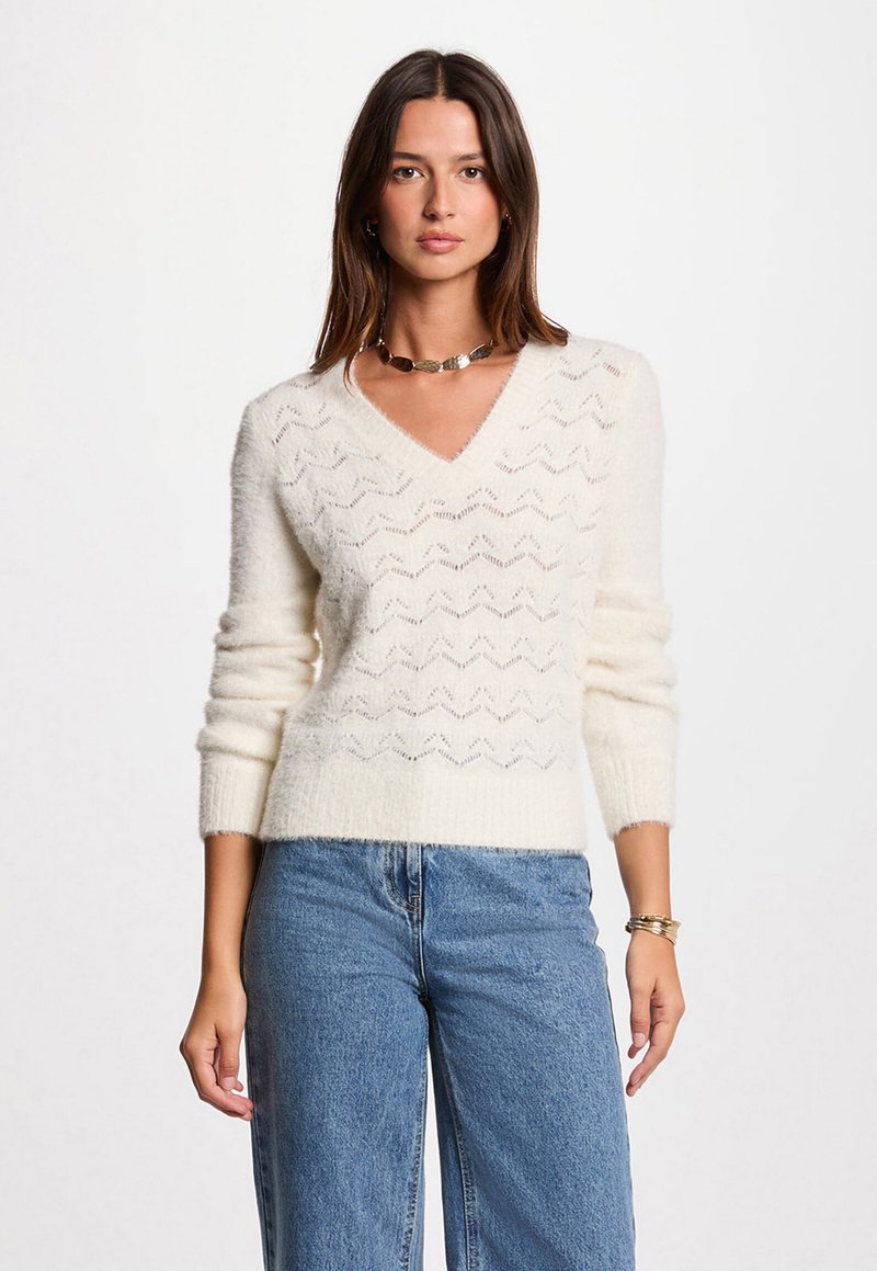 Weißer V-Ausschnitt-Pullover mit einem Zickzack-Strickmuster, langen Ärmeln, weichem Material, kombiniert mit hoch taillierten blauen Jeans.