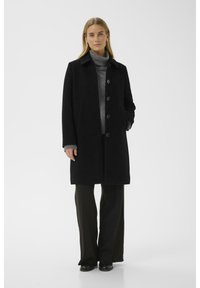 Schwarzer Mantel mit Kragen, Knopfleiste und seitlichen Taschen. Getragen über einem grauen Rollkragenpullover. Die gerade geschnittenen Hosen und schwarze Schuhe runden den Look ab.