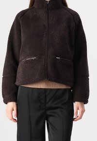 Giacca di pile marrone con colletto alto, chiusura frontale con zip e due tasche con zip; orlo e polsini a coste; texture morbida.