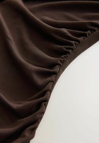 Ruskea venyvä kangas, jossa on kerrostunut rakenne, tunnusomaista sileä pinta ja pehmeä laskos. Kaareva reuna, jossa on minimaalinen ompeluyksityiskohta.