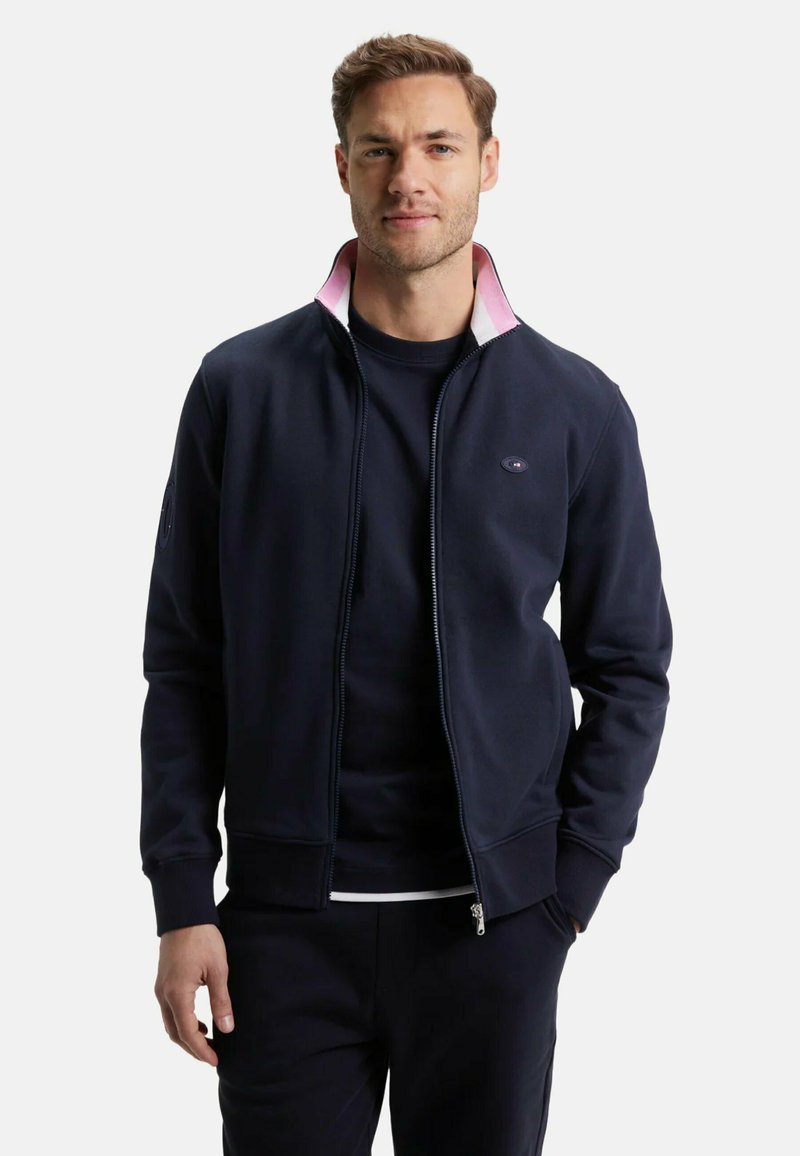 Veste zippée navy à col haut, avec une doublure intérieure rose, des poignets côtelés et un logo sur la poitrine. Texture douce et lisse.