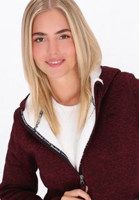 Burgundijas krāsas zip-up kapuce ar teksturētu audumu un baltu, pūkainu oderi. Iezīmes augsts apkakle un savilcējs, kontrastējoši iekšējie detaļas.