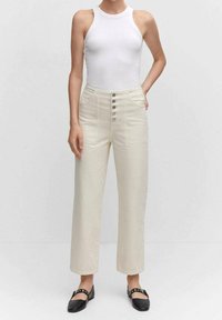 Pantalones beige de cintura alta con un diseño de pierna recta, que cuentan con cinco cierres de botón y bolsillos laterales. Llevados con una camiseta ajustada blanca.