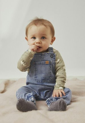 Jeans-Trägerhose mit einer Fronttasche, getragen über einem langärmligen, tiermusternden Hemd. Graue flauschige Socken auf einem strukturierten Deckenhintergrund.