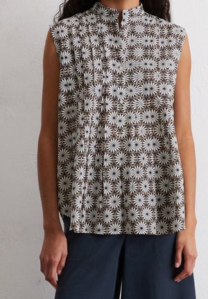 Bezrękawowa bluzka na guziki z geometrycznym kwiatowym wzorem w kolorach czarnym, białym i brązowym, noszona z ciemnymi spodniami.