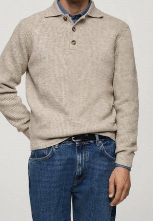 Hombre con suéter de punto beige con tres botones marrones sobre una camisa azul con cuello, combinado con vaqueros azules y cinturón negro.