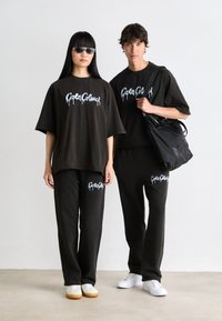 To personer står iført matchende svarte oversized skjorter og bukser med hvite logoer, hvite sneakers; en holder en svart veske over skulderen.