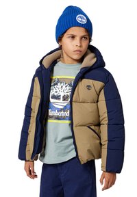 Gevoerde jas in kaki en marineblauw met een met bont gevoerde capuchon, gedragen over een lichtgroen shirt met een grafisch ontwerp en een blauwe gebreide beanie.