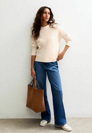 Pull en maille beige clair, jeans bleus évasés et sac fourre-tout marron. Baskets blanches avec des accents beiges. Textures lisses et coupe décontractée.