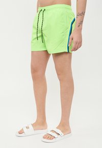 Pantaloni da bagno verde lime con elastico in vita e lacci neri. Presentano strisce laterali blu e nere. Indossati con ciabatte bianche.