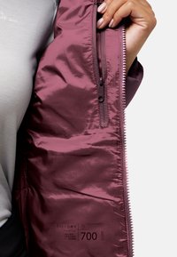 Una mano tiene aperto un giubbotto bordeaux che mostra una tasca con zip interna e il testo stampato "ExpeDRY 700 Allied Feather + Down" sul rivestimento.