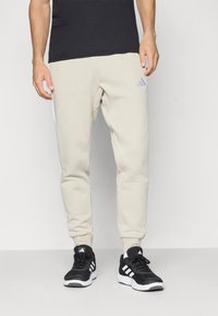 Vyras vilki šviesiai rudus Adidas treningus ir juodas Adidas sportbačius, stovintis prieš paprastą šviesų foną.