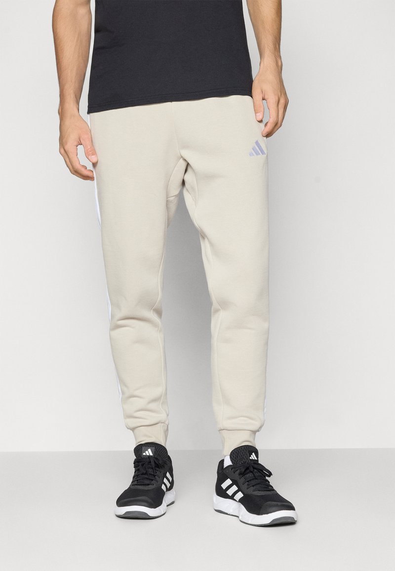 Vyras vilki šviesiai rudus Adidas treningus ir juodas Adidas sportbačius, stovintis prieš paprastą šviesų foną.