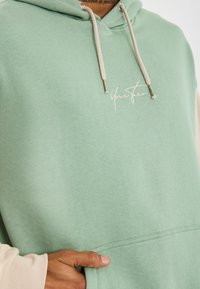 Persona con una felpa verde chiaro con lacci beige e la scritta "Il tuo turno" ricamata sul petto, mano appoggiata nella tasca frontale.