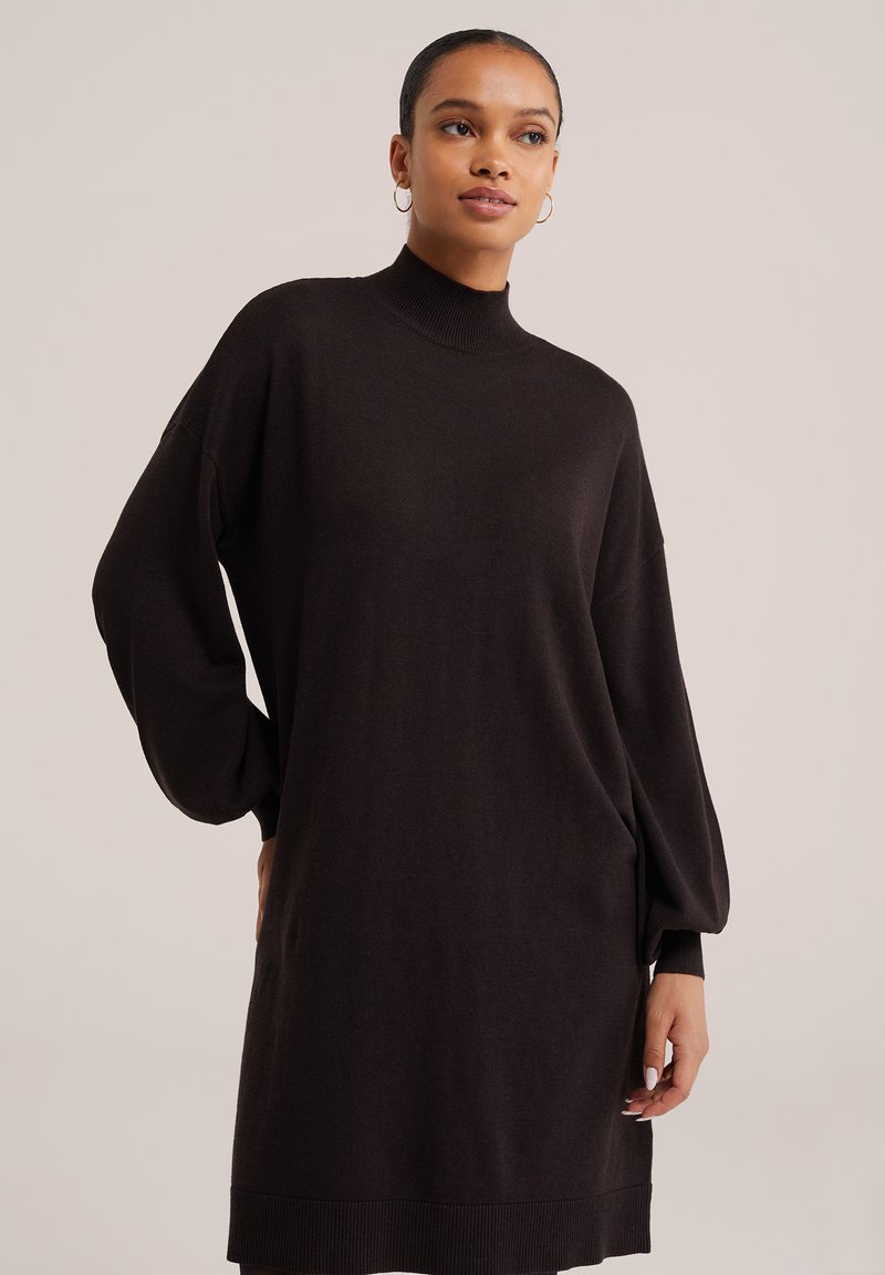 Femme portant une robe-pull noire à col roulé et manches longues, avec des boucles d'oreilles anneaux, debout devant un fond beige uni.