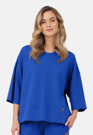 Kobieta o falowanych blond włosach, ubrana w luźny, jaskrawo niebieski, oversize'owy top i pasujące spodnie, stojąca na tle jednolitego jasnego tła.