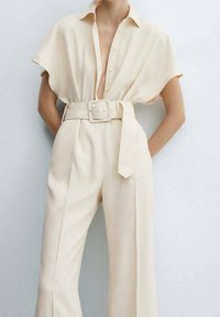 Crèmekleurige jumpsuit met riem, brede broekspijpen, korte mouwen en een diepe V-halslijn met knopen, gedragen door een staand persoon.