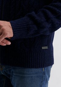 Dunkelblauer gestrickter Cardigan mit gerippten Bündchen und Saum. Merkmale ein strukturiertes Kabelmuster und ein kleines Markenschild an der Seite. Mit Reißverschluss.