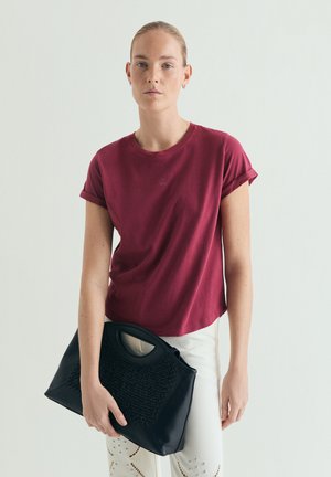 Jeune femme aux cheveux blonds portant un t-shirt bordeaux et un pantalon blanc, tenant un grand sac à main noir texturé sur un fond uni.