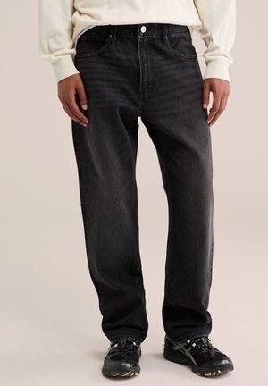 Person trägt lockere schwarze Jeans, ein weißes langärmeliges Hemd und schwarze Sportschuhe und steht auf hellem Hintergrund.
