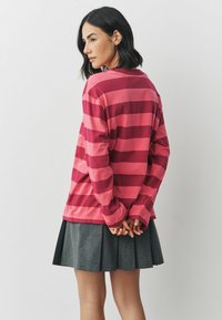 Frau mit kurzen dunklen Haaren, die ein langärmeliges, rosa-rot gestreiftes Shirt und einen grauen, plissierten Rock trägt, steht mit hinter dem Rücken gefalteten Händen.