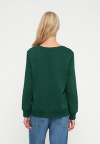 Sudadera verde oscura con un cuello amplio, mangas largas y puños de canalé; combinada con jeans de tiro alto en azul claro.