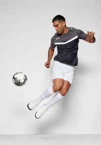 Schwarzes und graues Sportshirt mit weißem Streifen, kombiniert mit weißen Shorts und Socken. Person springt mit einem schwarz-weißen Fußball in der Nähe.