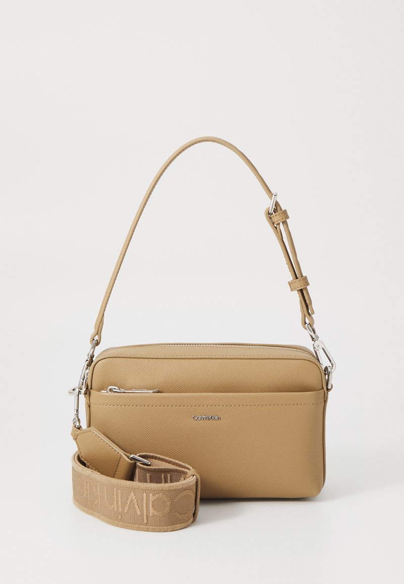 Tan crossbody bag with textured faux leather, slim rectangular shape, top zip closure, and a woven branded strap with metal hardware.