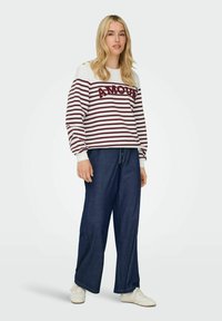 Sweatshirt rayé en blanc et bordeaux avec le texte "AMOUR", associé à un pantalon large bleu marine et des baskets blanches. Détails de boutons visibles.