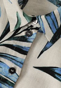Lys beige skjorte med farverigt bladmønster i grønt, blåt og sort. Har sorte knapper og en krave-design med et struktureret stof.