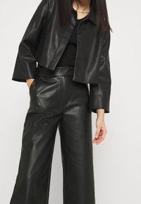 Giacca in pelle nera corta con colletto, abbinata a pantaloni larghi neri in ecopelle a vita alta. Texture liscia e chiusura con bottone.