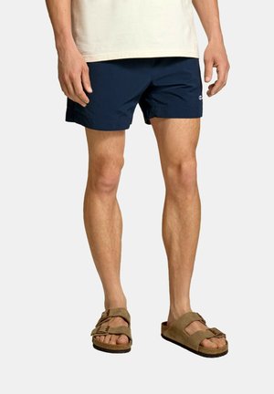 Mand iført marineblå shorts, beige t-shirt og brune ruskind dobbeltsnøresandaler står mod en ensfarvet baggrund.