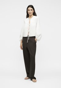 Γυναίκα όρθια, φοράει άσπρο μακρυμάνικο μπλουζάκι με ζώνη μπροστά, μαύρο παντελόνι με φαρδιά μπατζάκια και μπεζ φλιπ-φλοπς σε λευκό φόντο.