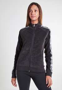 Giacca in pile nera con zip e colletto alto, caratterizzata da strisce laterali bianche e polsini a coste. Tessuto morbido, design aderente.