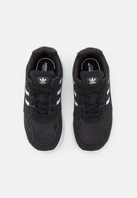 Un paio di sneaker Adidas nere con strisce bianche e suole bianche, viste dall'alto su uno sfondo grigio chiaro.