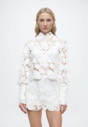 Žena v bielom kvetinovom čipkovanom topu s dlhými rukávmi s nadýchanými rukávmi a rovnako čipkovaných šortkách na jednoduchom pozadí.