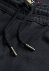 Close-up van zwarte stoffen broek met zwarte koordjes featuring metalen uiteinden gegraveerd met "SUPERDRY."