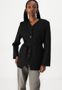 Blazer noir avec un col en V, boutons à l'avant, longues manches et taille ceinturée. Tissu lisse avec deux poches latérales et coupe ajustée.