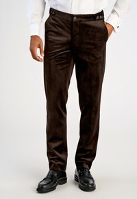 Mand iført mørkebrune slim-fit ruskindsbukser med sidetilpasningsknapper, parret med sorte læder loafers og en hvid skjorte.