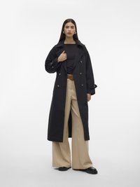 Femeie purtând o carwear lungă neagră peste un top negru, pantaloni bej cu talie înaltă și curea maro, pantofi negri, stând în picioare pe fundal alb simplu.