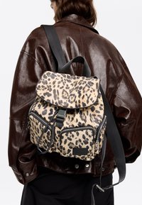 Plecak z tkaniny w panterkę z klapką na górze, dwoma przednimi kieszeniami na suwak oraz regulowanymi czarnymi paskami. Charakteryzuje się eleganckim, teksturowanym wzorem.