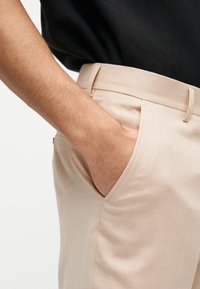 Χέρι στην μπροστινή τσέπη μπεζ παντελονιού που φοριέται με μαύρο πουκάμισο, παρουσιάζοντας την υφή του υφάσματος και τις θηλιές της ζώνης.