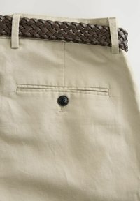 Pantalones de tela beige con un cinturón de cuero trenzado marrón oscuro. Presenta un bolsillo trasero con cierre de botón negro y una superficie texturada sutil.