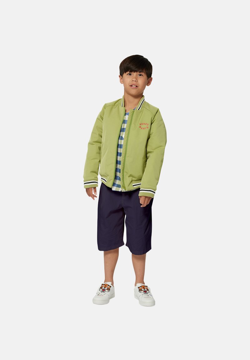 Groene bomberjack met rood "KENZO PARIS" logo, blauwe plaid overhemden, marineblauwe shorts en witte sneakers met tijgerprint accenten.