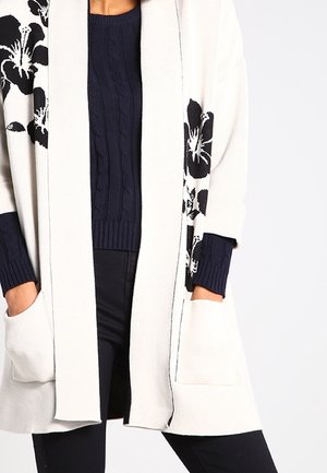 Cardigan crème à ouverture avant avec de grands motifs floraux noirs sur les manches, porté sur un pull en maille torsadée bleu marine et un pantalon noir.